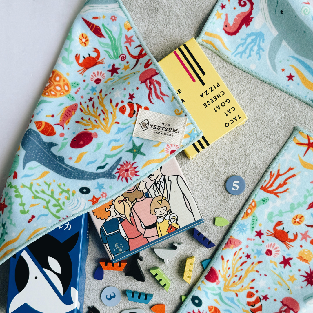 Half opened Tsutsumi Clingy Wrap with marine creatures artwork on light blue background, holding small boxes of games and game tokens.