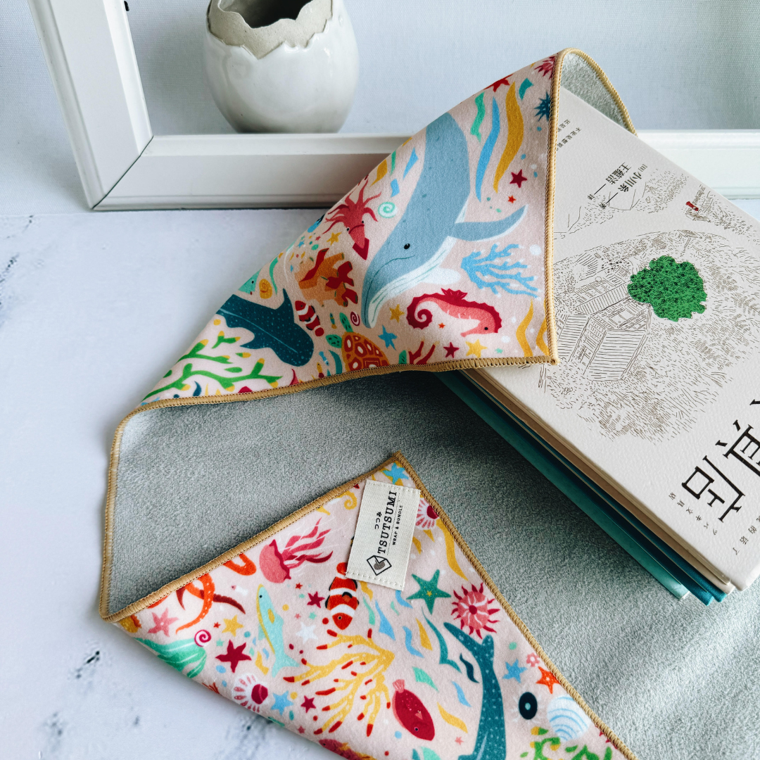 Half opened Tsutsumi Clingy Wrap with marine creatures artwork on light pink background, wrapping some books.