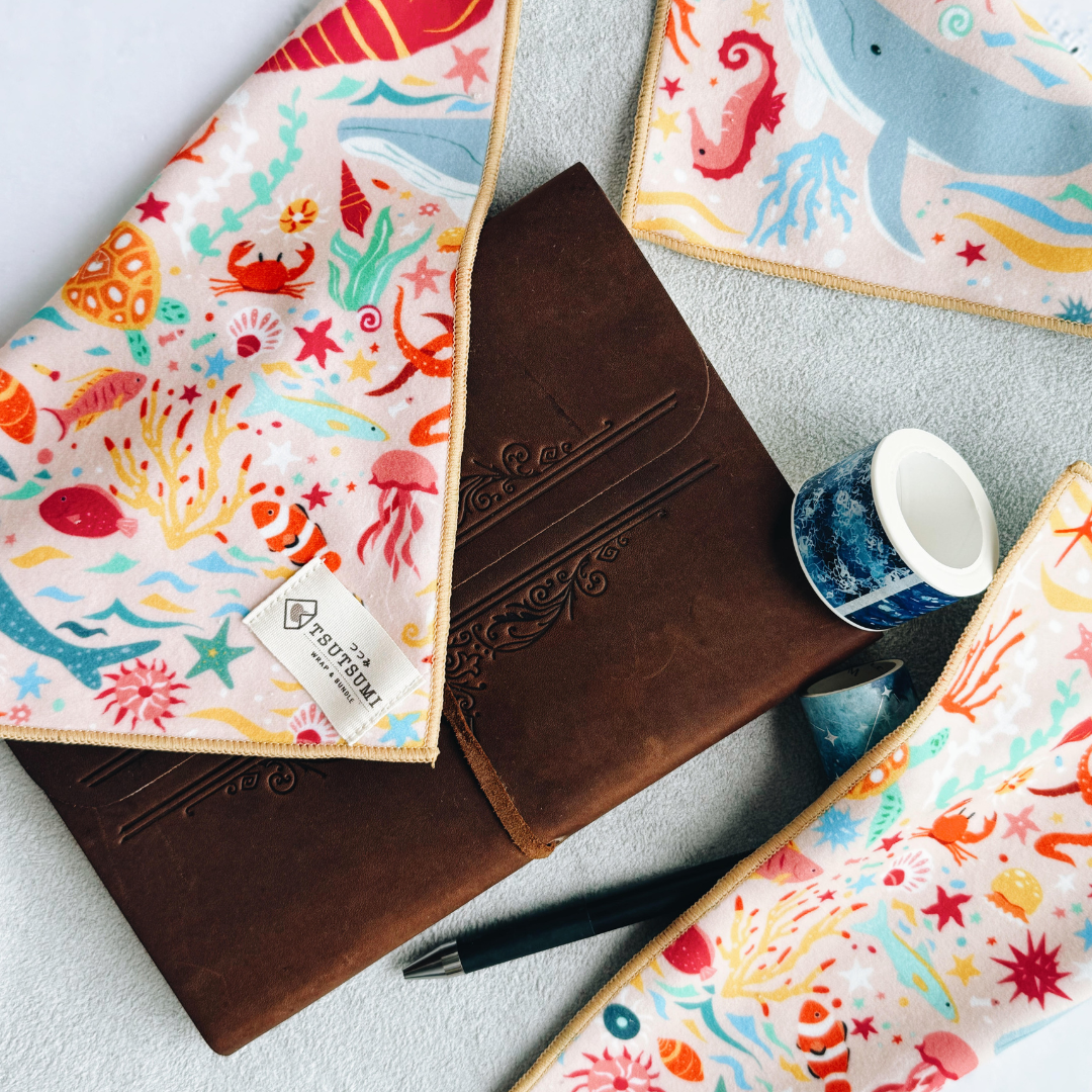 Half opened Tsutsumi Clingy Wrap with marine creatures artwork on light pink background, wrapping a journal notebook, tapes and pen.