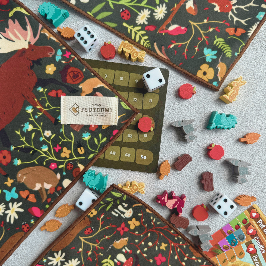 Half opened Tsutsumi Clingy Wrap with woodland creatures artwork and dark green background used to wrap some board game tokens, dies and cards.
