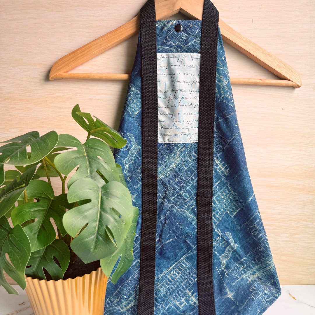 A blue patterned fabric wrap with black straps hanging on a wooden hanger against a light wood wall, with a green potted plant in the foreground.