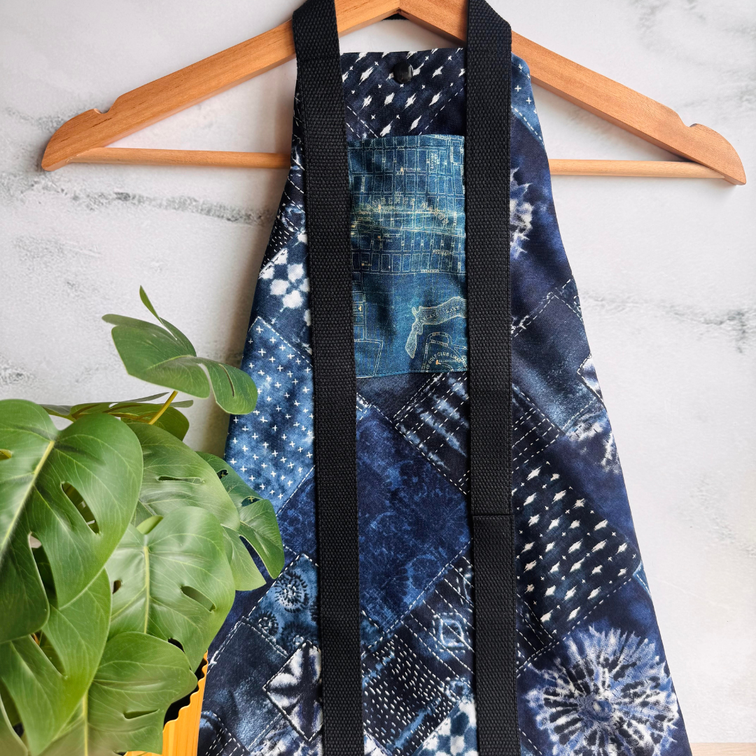 Blue Sashiko Patchwork patterned fabric wrap with black straps on a wooden hanger against a white background with a plant.