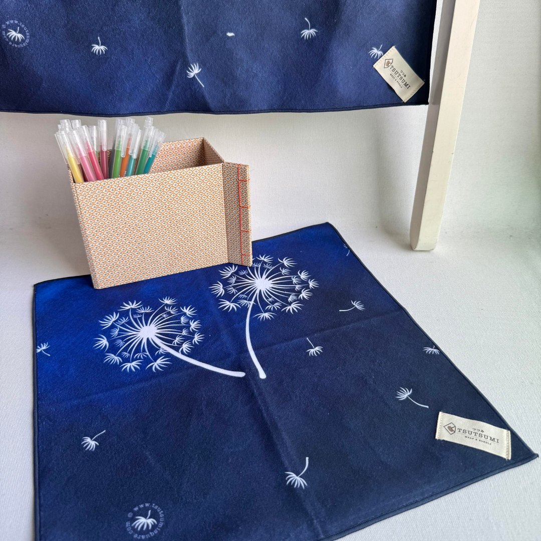 Two blue fabric squares with white dandelion designs, with a box of colorful pens