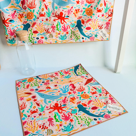 A big piece of fabric with Marine Fantasy print in light pink background hanging from a rack and a small piece of fabric with Marine Fantasy print in light pink background laying on the table.