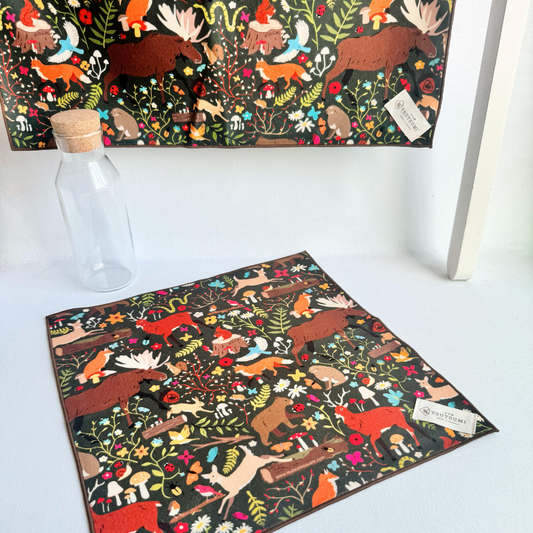 Fantasy print in dark green background hanging from a rack and a small piece of fabric with Woodland Fantasy print in dark green background laying on the table