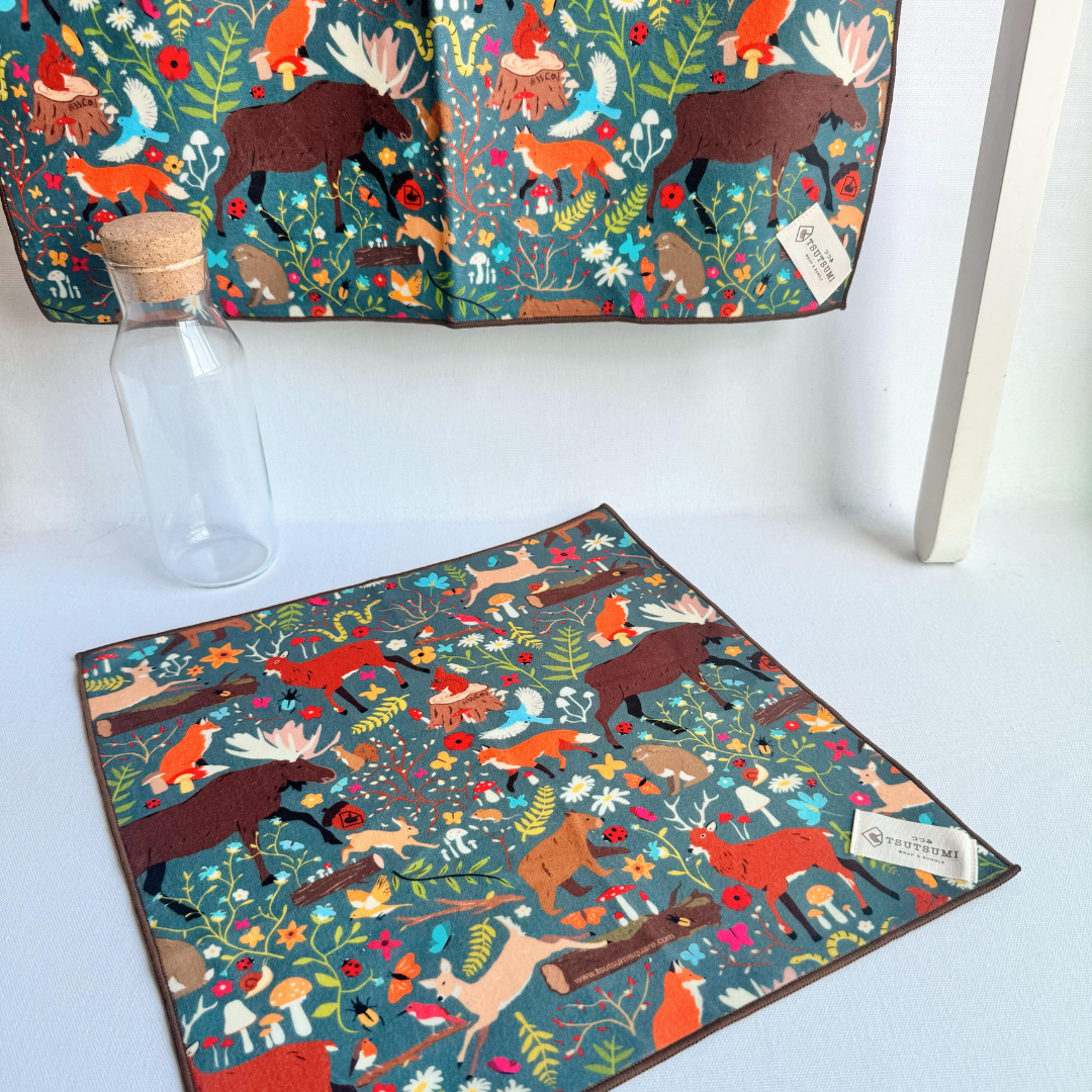 a big piece of fabric with Woodland Fantasy print in dark teal background hanging from a rack and a small piece of fabric with Woodland Fantasy print in dark teal background laying on the table