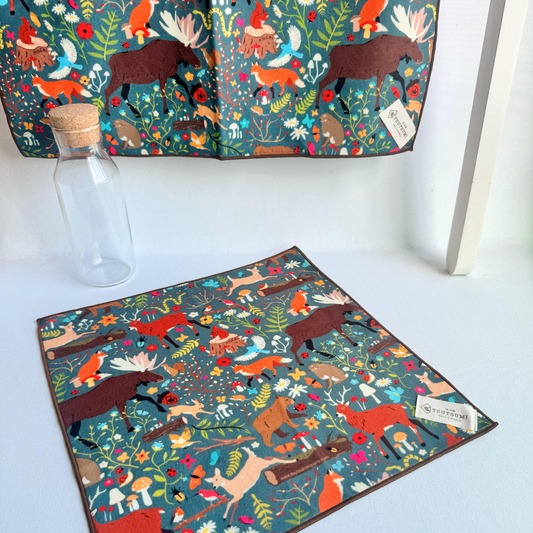 a big piece of fabric with Woodland Fantasy print in dark teal background hanging from a rack and a small piece of fabric with Woodland Fantasy print in dark teal background laying on the table
