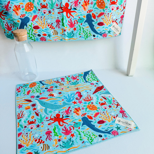 A big piece of fabric with Marine Fantasy print in light blue background hanging from a rack and a small piece of fabric with Marine Fantasy print in light blue background laying on the table.