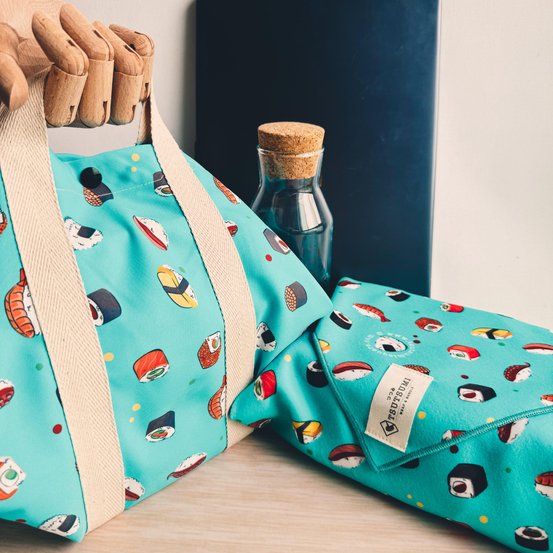 2 pieces of blue fabric with sushi pattern, a square wrap with handles wrapping a lunchbox, and a clingy wrap with Tsutsumi brand label wrapping some books. 