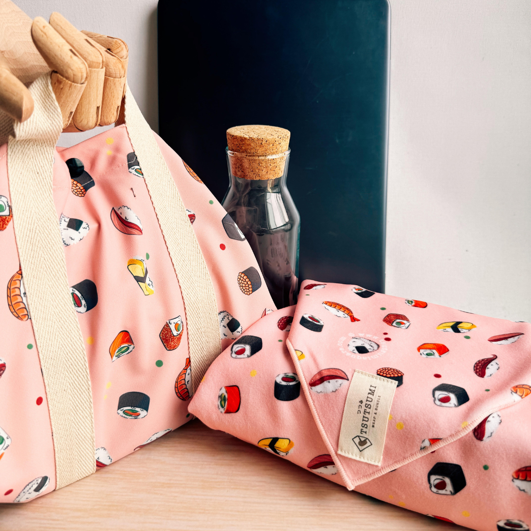 2 pieces of Pink fabric with sushi pattern, a square wrap with handles wrapping a lunchbox, and a clingy wrap with Tsutsumi brand label wrapping some books. 