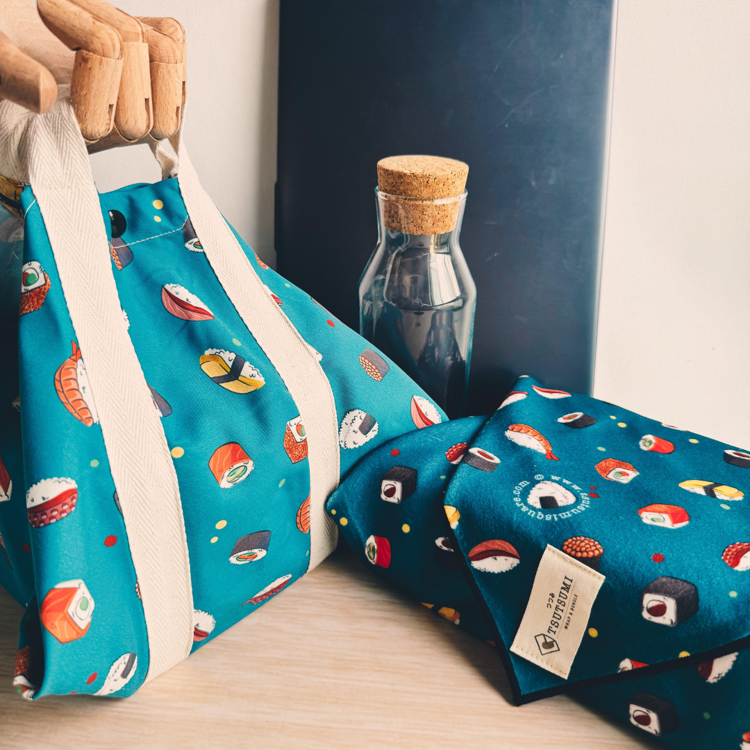 2 pieces of Dark Teal fabric with sushi pattern, a square wrap with handles wrapping a lunchbox, and a clingy wrap with Tsutsumi brand label wrapping some books. 