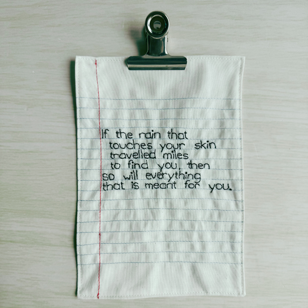 Fabric notebook page with a handstitched quote and a binder clip on a wooden surface