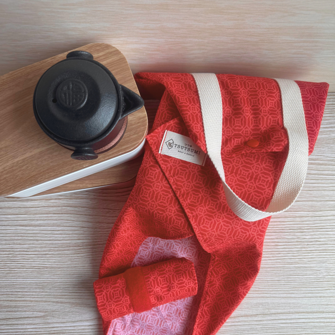 Red patterned fabric carrier with a white label on a wooden surface next to a black tea pot.