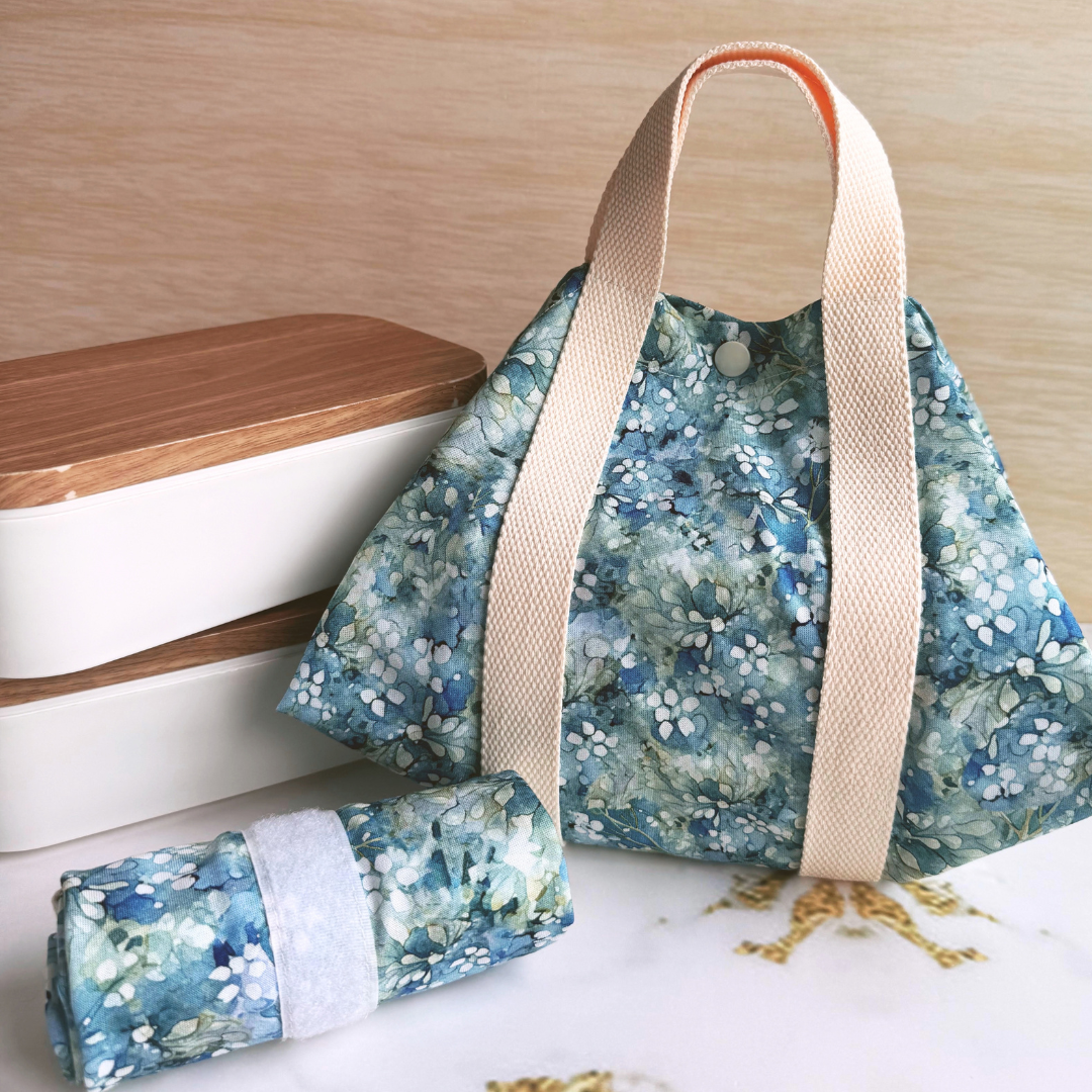 A reusable fabric tote bag with a blue and green floral print, beige cotton handles, and a matching rolled-up pouch placed in front, set on a tabletop with stacked containers in the background.