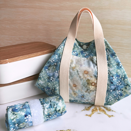 A fabric wrap with a blue and white floral print, beige handles, and a matching rolled-up pouch placed beside it on a light surface against a wood-toned background.