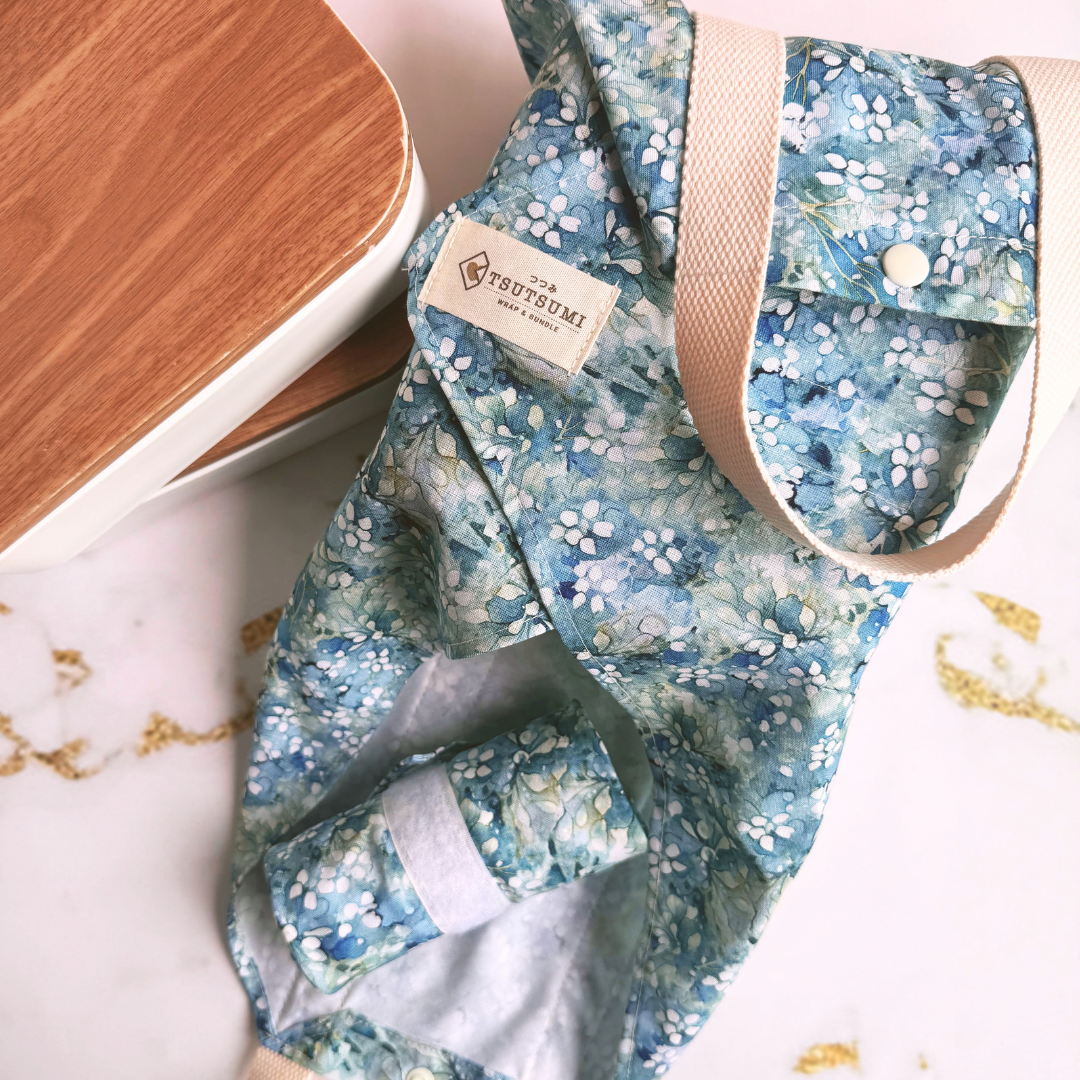 A fabric wrap with a blue and white floral print, beige handles, and a matching rolled-up pouch placed beside it on a light surface against a wood-toned background.