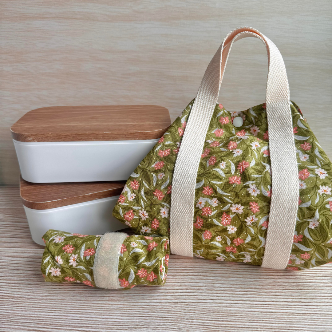 Green floral-patterned fabric square wrap with beige strap handles, next to two lunchboxes on a wooden surface