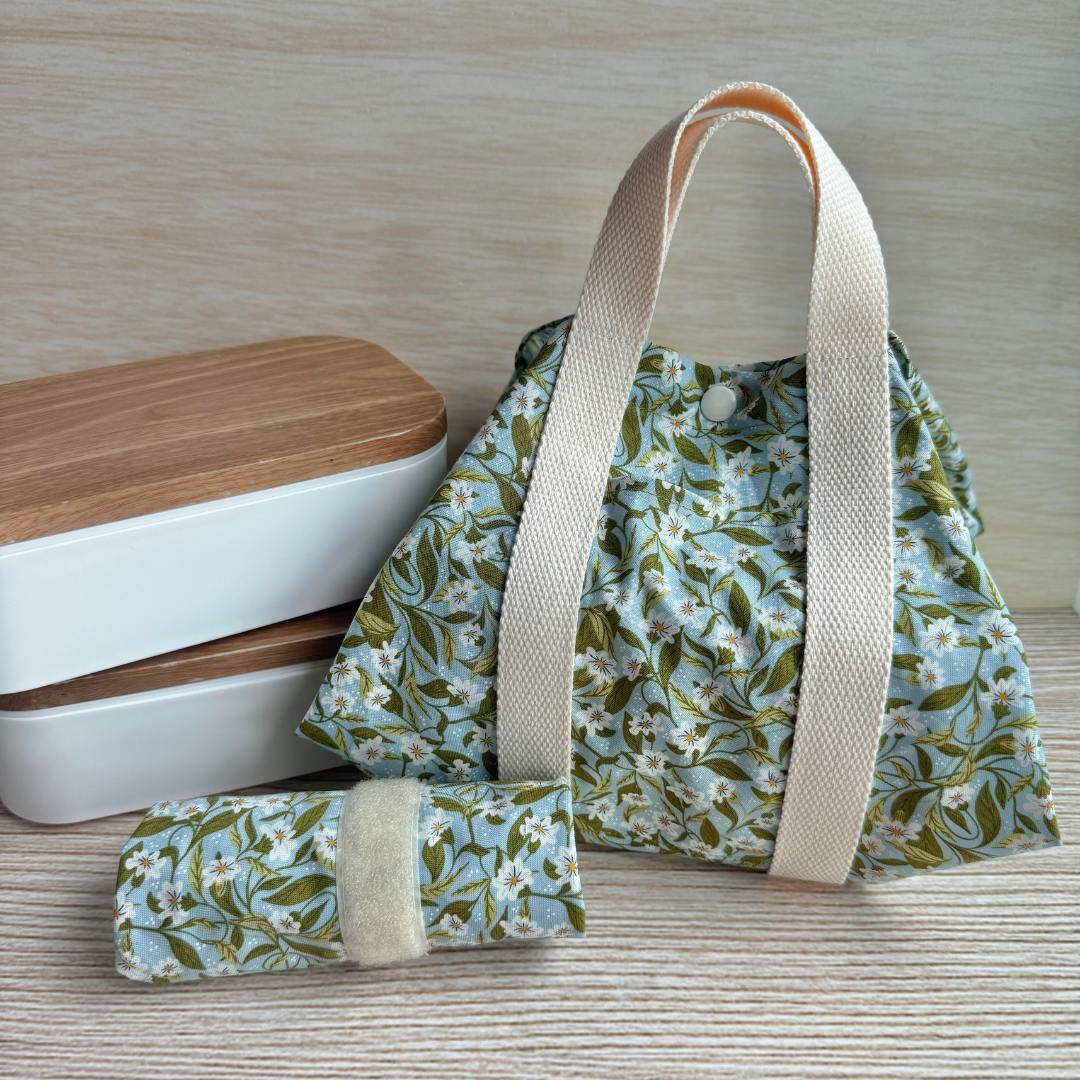 Floral-patterned fabric square wrap with beige handle straps next to 2 lunchboxes