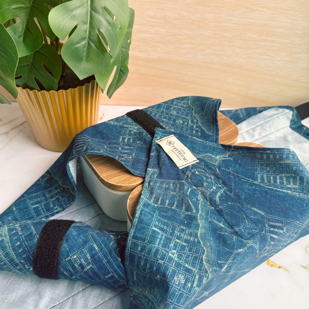A blue patterned fabric pouch or wrap with black velcro closures and a small sewn label, shown folded on a light surface next to a potted green plant and a gold planter.