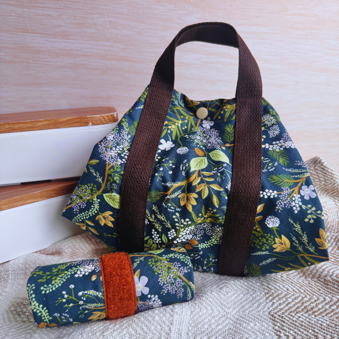 A reusable fabric wrap with a dark green floral print, brown handles, on a light fabric surface beside two lunch boxes.