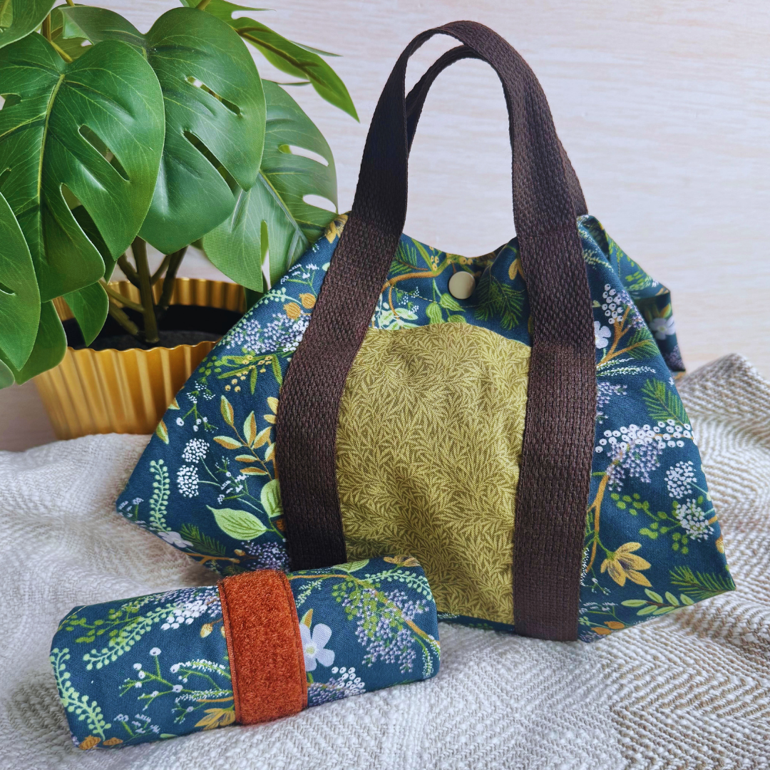 A reusable fabric wrap with a dark green floral print, brown handles, and a textured olive-green front panel, shown with a matching rolled-up wrap on a light fabric surface beside a potted green plant.