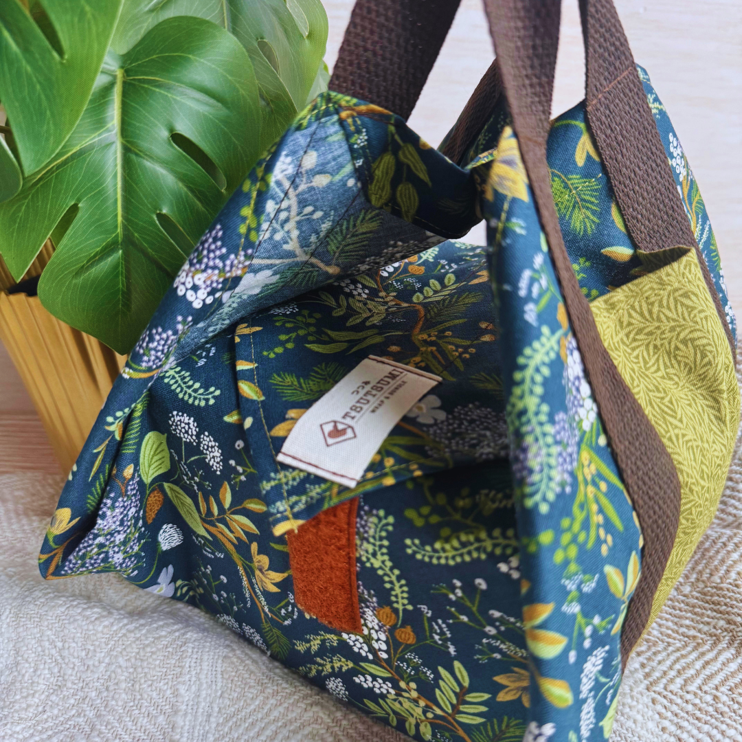 A reusable fabric wrap with a dark green floral print, brown handles and a brand label, on a light fabric surface beside a pot of green plant.