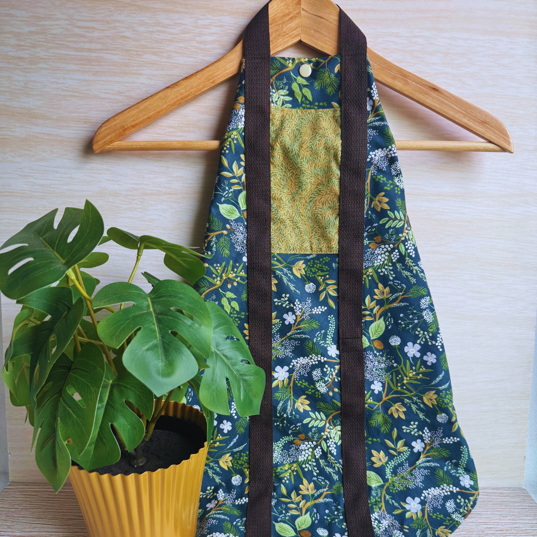 A reusable fabric wrap with a dark green floral print, brown handles, and a textured olive-green front panel, shown hanging from a clothes hanger beside a potted green plant.