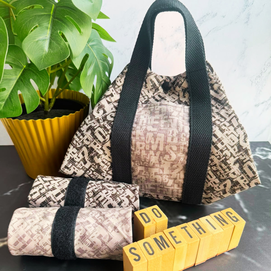 A black patterned fabric square wrap with black handles sits on a dark tabletop next to a potted green plant. In front of the square wrap are two matching rolled fabric items with black bands, and yellow letter blocks spell out 'DO SOMETHING'.