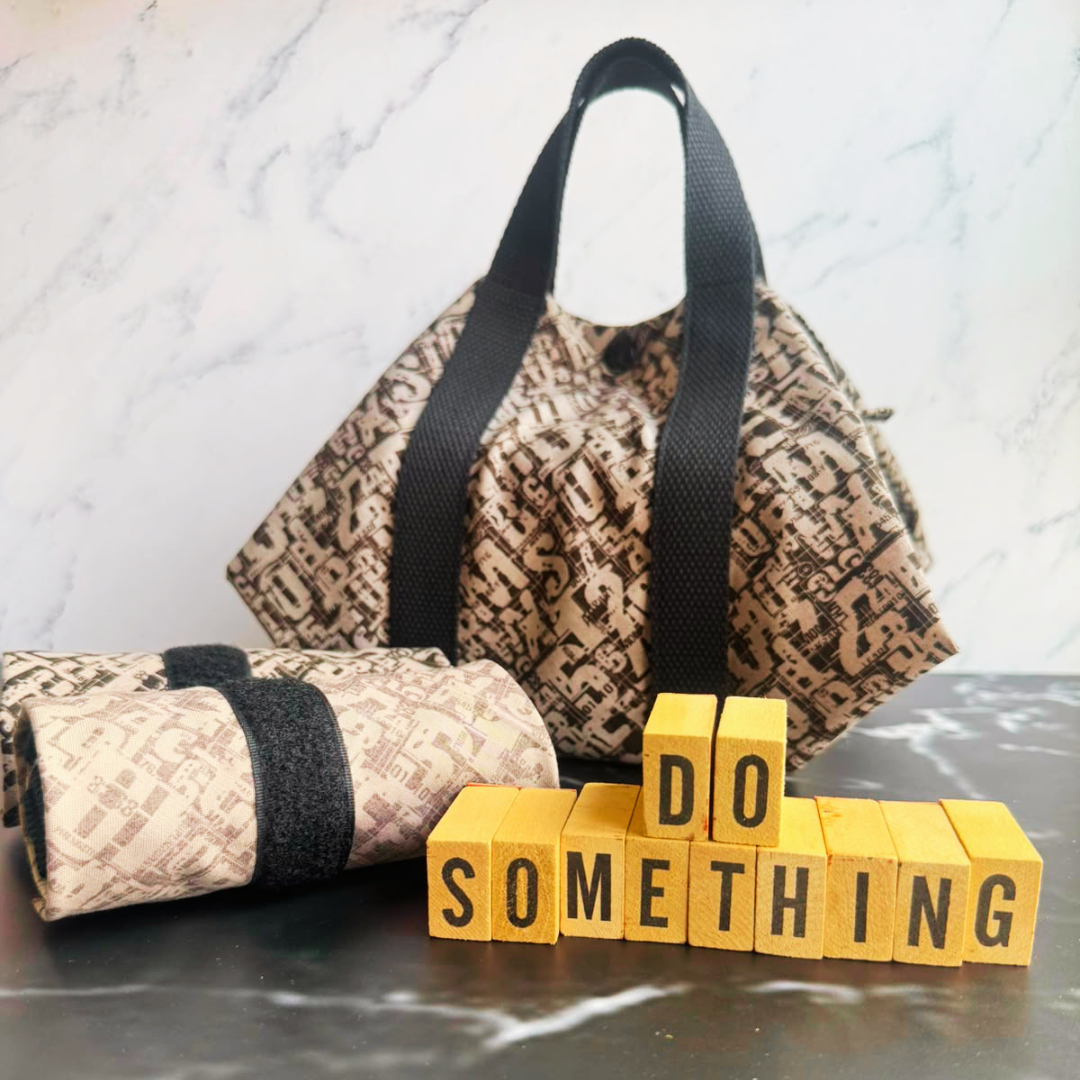 A black patterned fabric square wrap with black handles sits on a dark marble-like surface, with a matching rolled fabric item in front and yellow wooden blocks spelling 'DO SOMETHING' in front of it. The background is a light marble wall.