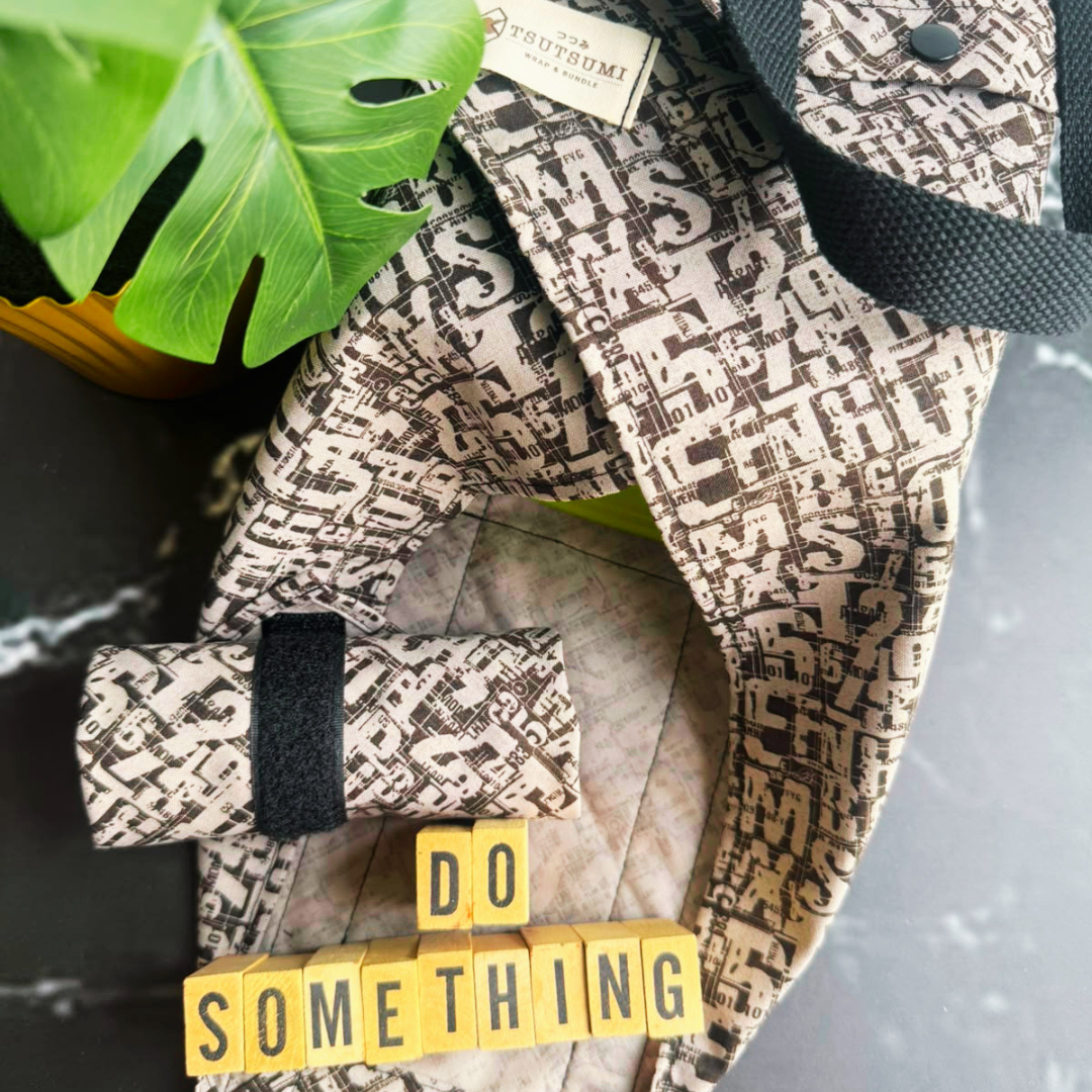 A close-up of a black patterned fabric square wrap with a matching rolled wrap, styled with a green monstera leaf, yellow letter blocks spelling 'DO SOMETHING', and a dark marble surface.