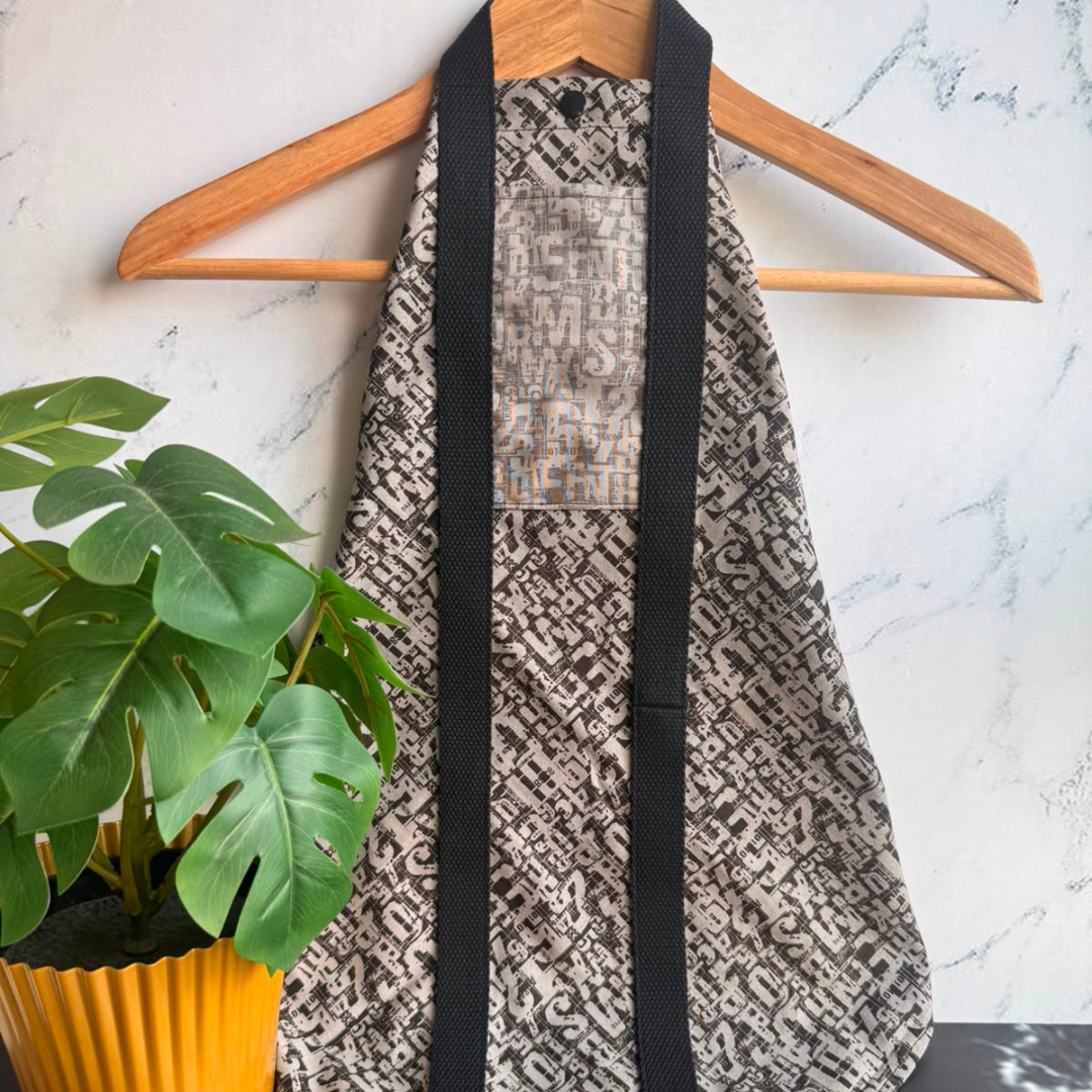 A black patterned fabric square wrap with black straps hanging on a wooden hanger against a light marble wall, with a green potted plant in the foreground.