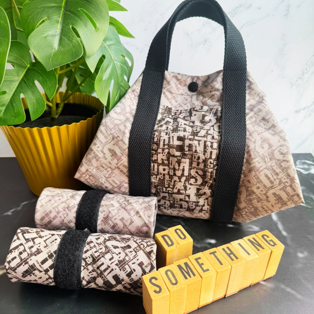 A light grey patterned fabric square wrap with black handles sits on a dark tabletop next to a potted green plant. In front of the square wrap are two matching rolled fabric items with black bands, and yellow letter blocks spell out 'DO SOMETHING'.