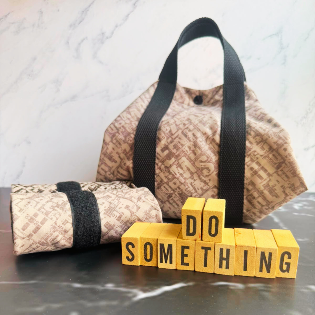 A light grey patterned fabric square wrap with black handles sits on a dark marble surface, with a matching rolled wrap in front and yellow letter blocks spelling 'DO SOMETHING' in the foreground.