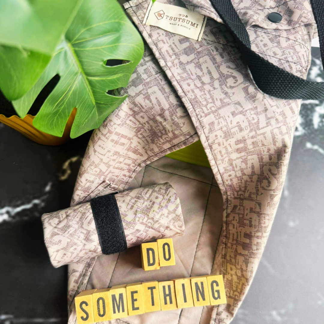 A close-up of a light grey patterned fabric square wrap with a matching rolled wrap, styled with a green monstera leaf, black marble surface, and yellow letter blocks spelling 'DO SOMETHING'. 
