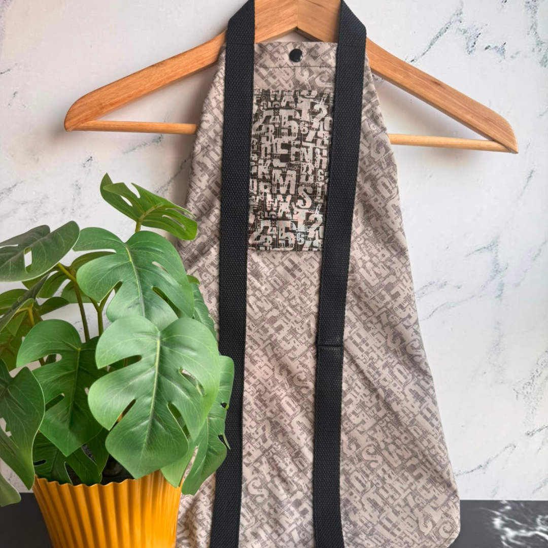A light grey patterned fabric square wrap with black straps hanging on a wooden hanger against a light marble wall, with a green potted plant in the foreground.