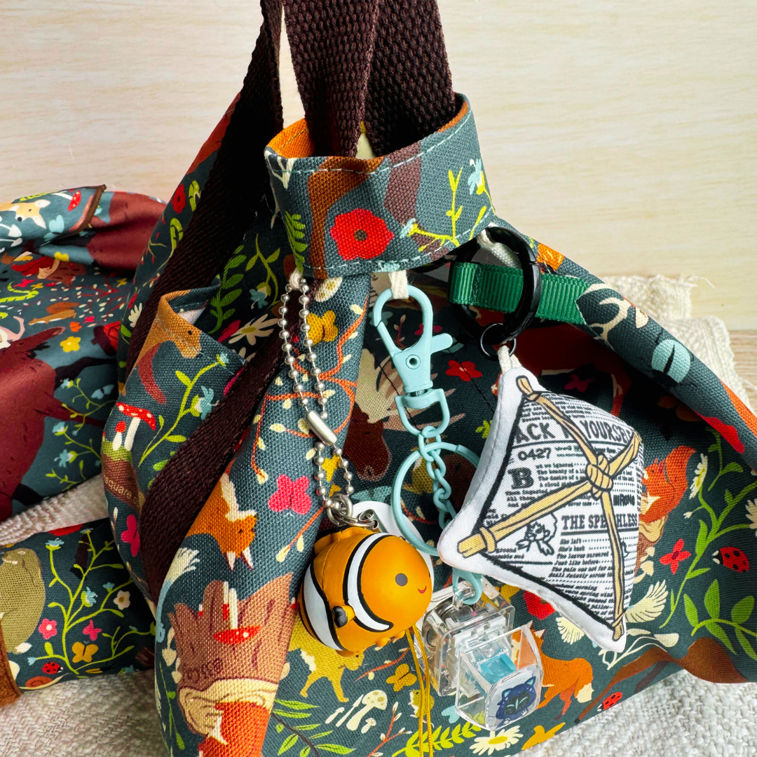 Keychains hanging from a strap attached to handles of Tsutsumi square wrap with woodland-themed pattern.