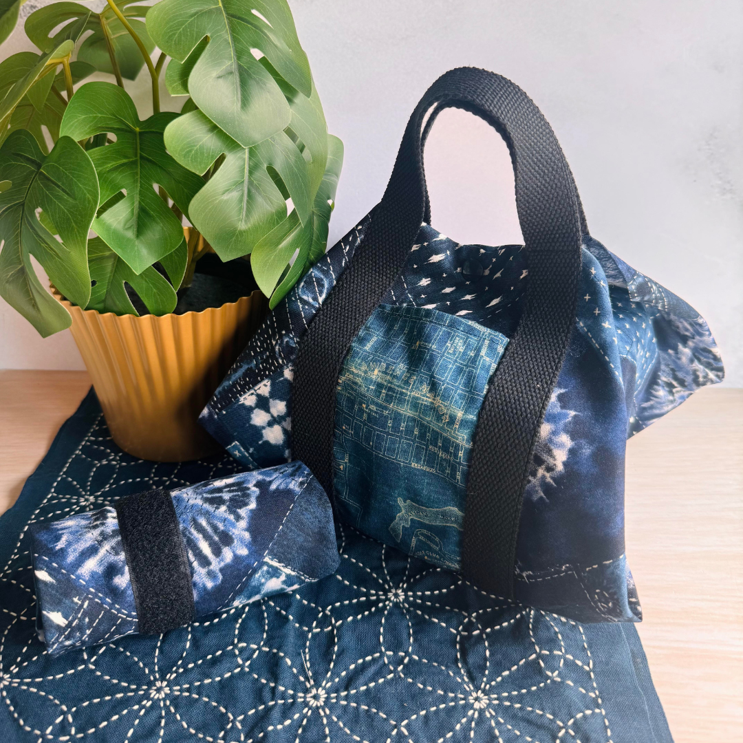 Blue Sashiko Patchwork patterned bag with black straps on a wooden surface next to a potted plant.
