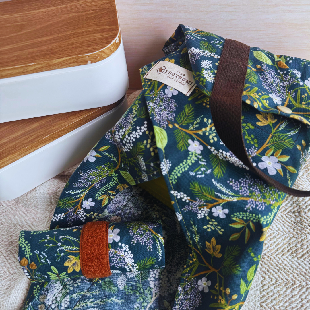 A reusable fabric wrap with a dark green floral print, brown handles, wrapping a lunch box, shown with a matching rolled-up wrap on a light fabric surface beside two lunch boxes.